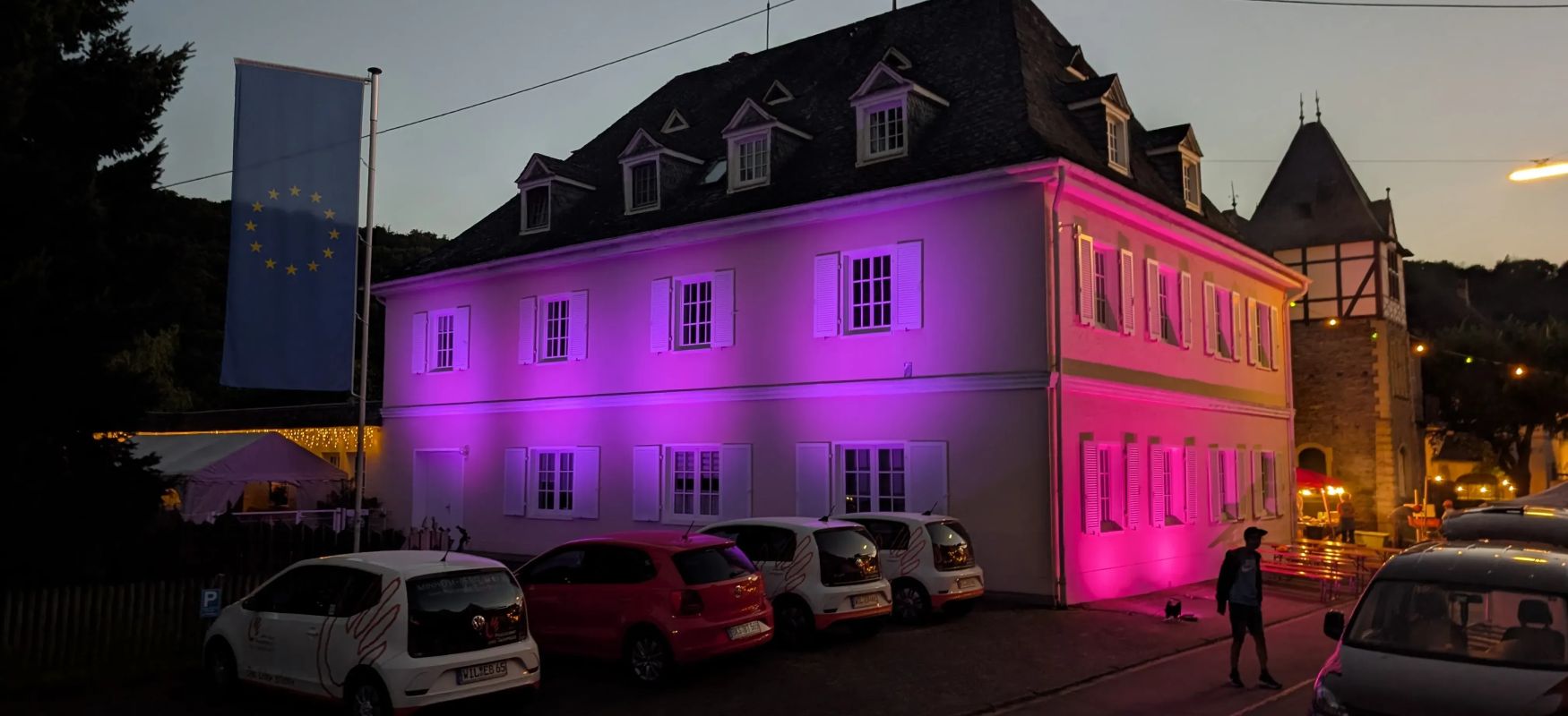 Weisses haus mit pinker beleuchtung mehrere autos davor europafahne am abend und ein mann spaziert