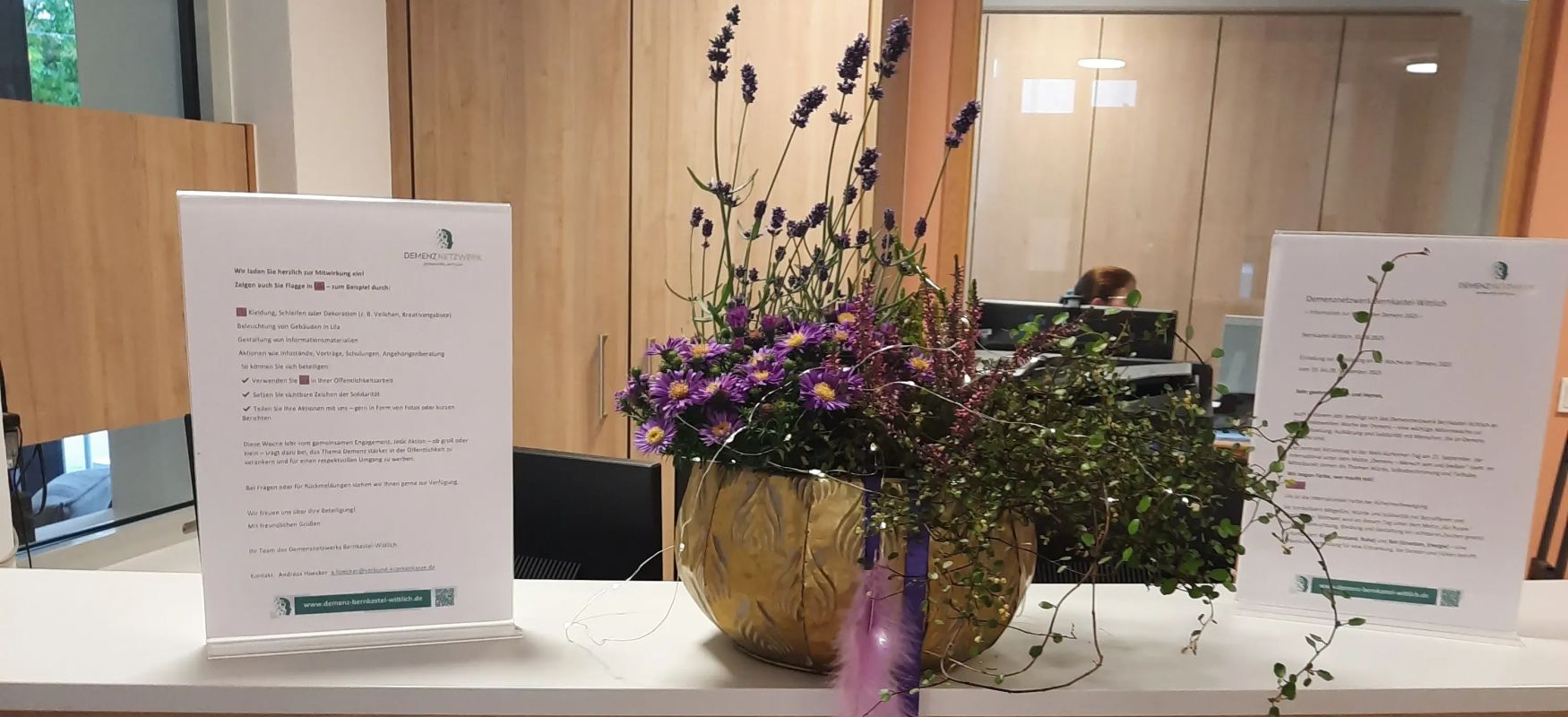 Blumenarrangement mit lila blumen auf empfangstheke mit holzdekor und zwei infoblättern im hintergrund