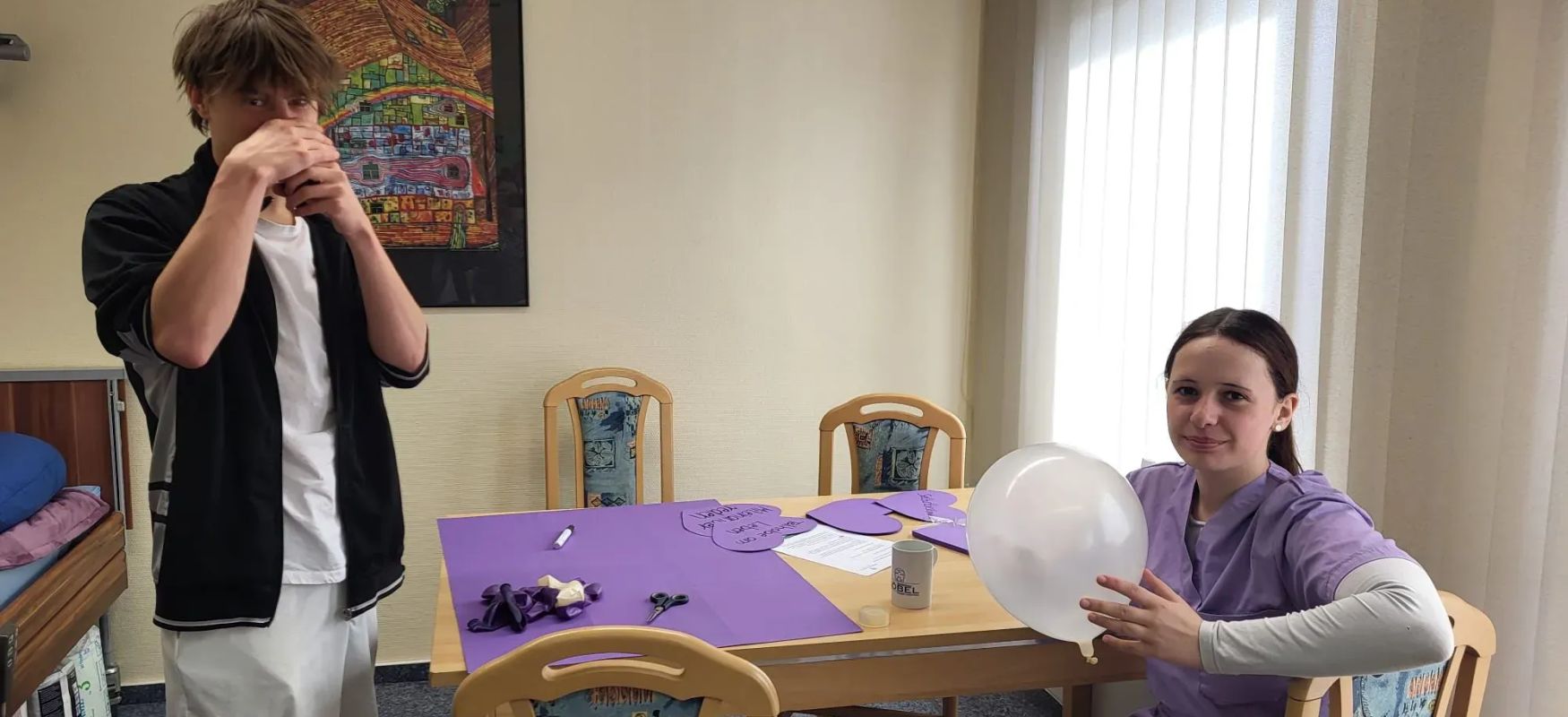 Zwei Jugendliche in einem Raum einer Pflegeeinrichtung Junge bläst Luftballon auf Mädchen sitzt am Tisch mit Bastelmaterialien
