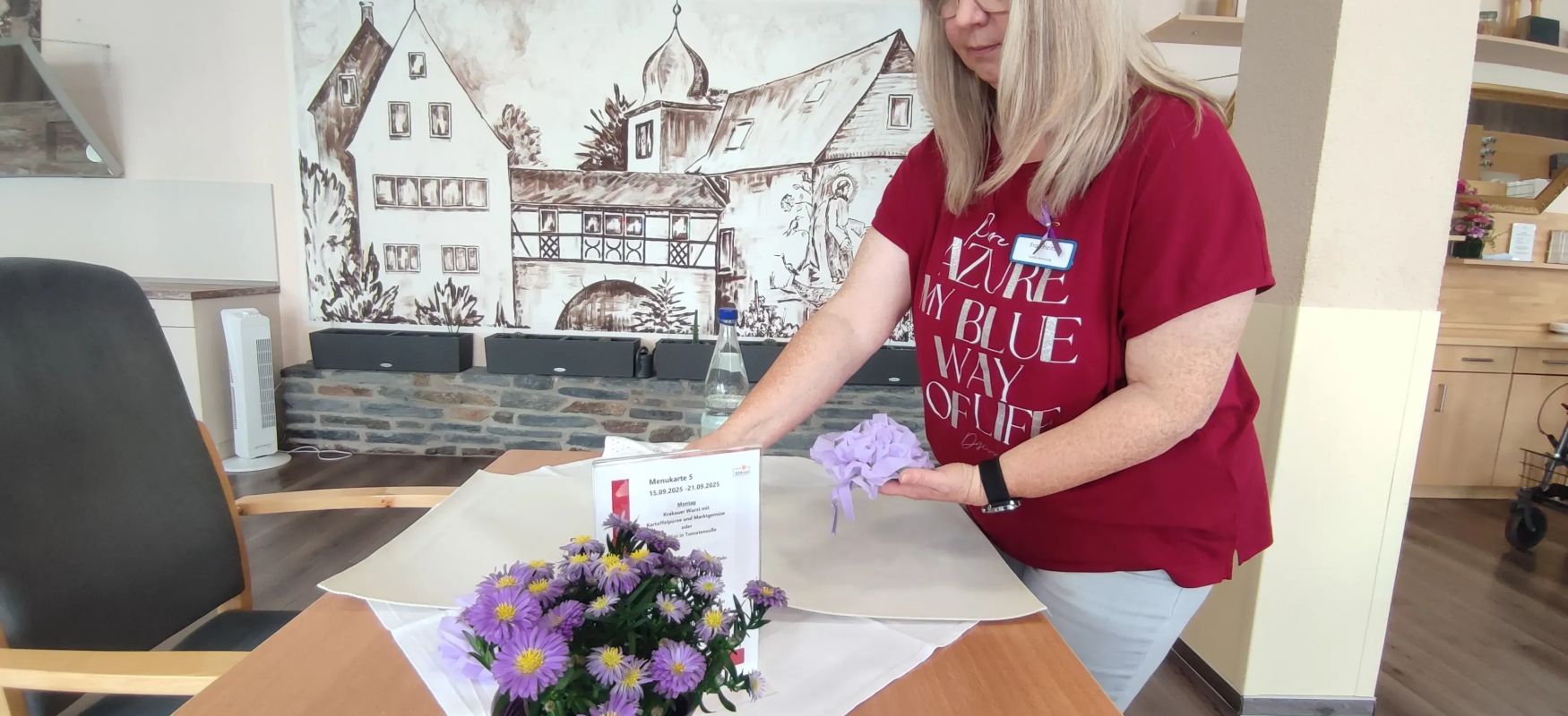 Frau mit rotem Shirt deckt Tisch in einem Raum mit Gemälde und lila Blumen auf dem Tisch
