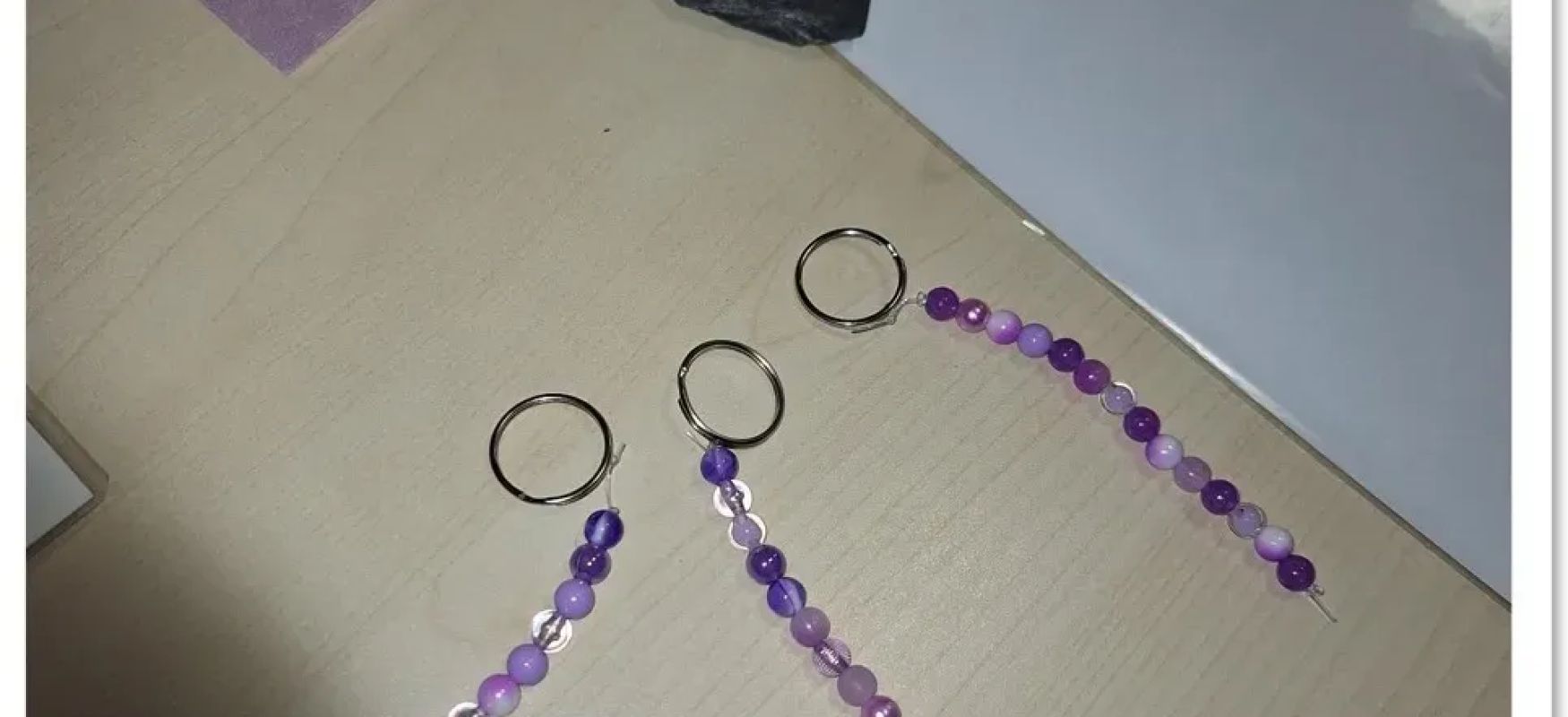 Drei lila Perlenanhänger mit metallringen auf hellem Holztisch und schwarzem stein mit Schrift