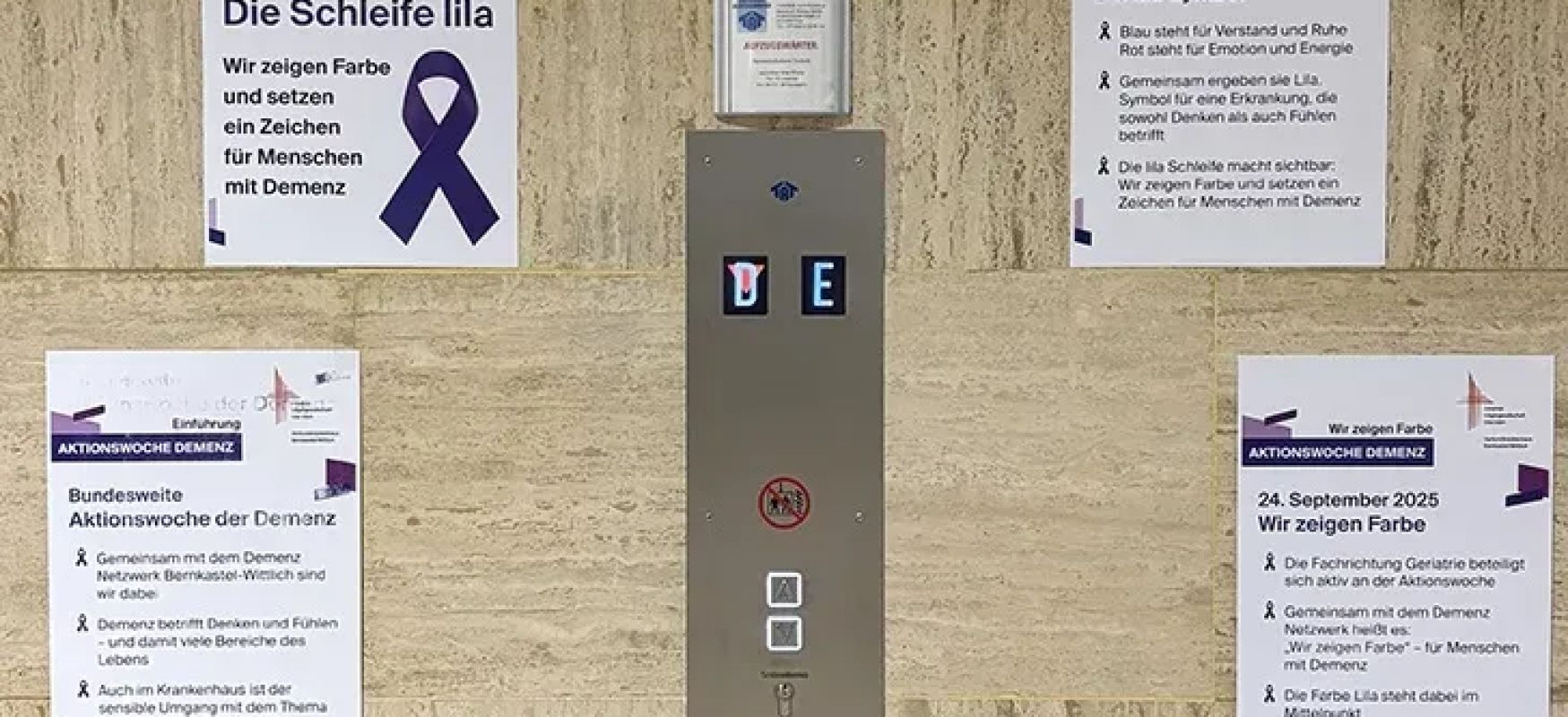 Plakate zur Aktionswoche Demenz mit lila Schleife an heller Wand neben Aufzugsbedienfeld aufgezogen auf deutsch