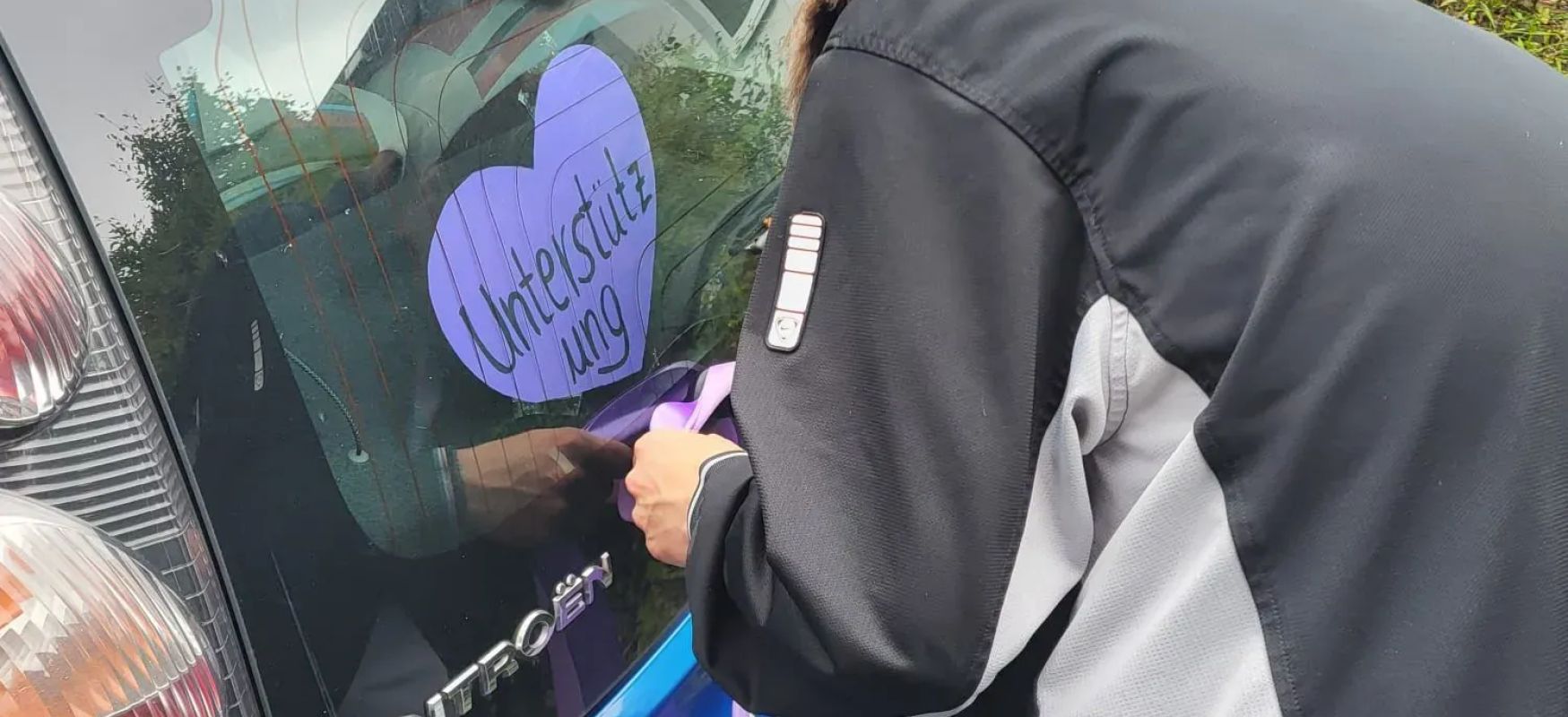 Person bindet lilafarbenes Band an blaue Ambulanzfahrzeug mit Herz Aufkleber und Schriftzug Unterstützung